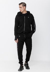 Czarna bluza z zamkiem błyskawicznym, z przednimi kieszeniami, dopasowane czarne dresy z wzorzystymi bocznymi paskami oraz czarne adidasy, tworzące swobodny wygląd.