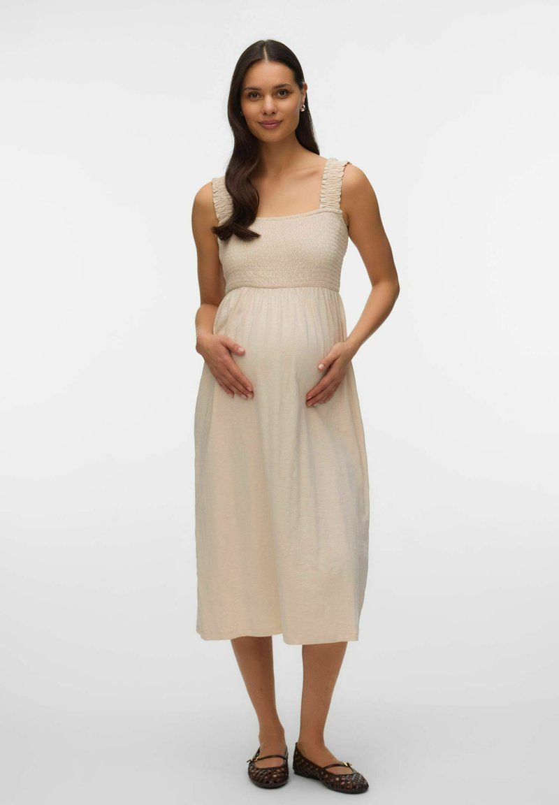 Femme enceinte aux longs cheveux foncés portant une robe midi beige sans manches et des ballerines tissées marron, tenant son ventre.