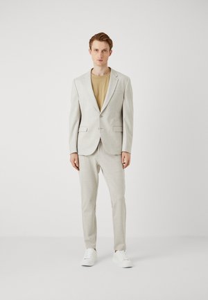 Lys grå dressjakke med slag og to knapper, kombinert med matchende bukser og en beige T-skjorte. Hvite sneakers fullfører antrekket.