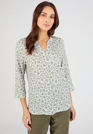 Frau trägt eine weiße Bluse mit grauem Blumenmuster und dreiviertellangen Ärmeln, kombiniert mit olivgrünen Hosen, steht vor einem einfarbigen Hintergrund.