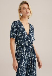 Marinblå jumpsuit med korta ärmar, djup V-ringning, knytbälte och vitt abstrakt mönster. Tillverkad av mjukt, texturerat tyg.