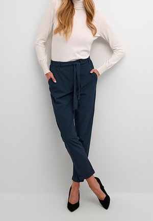 Navy trousers with a belted waist, tapered legs, and rolled cuffs, paired with a cream turtleneck and black high heels against a neutral backdrop.