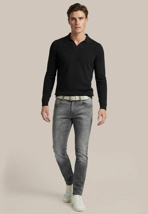 Mann geht nach vorne, trägt schwarzes langärmliges Polo, graue ausgewaschene Jeans, weißen Gürtel und weiße Turnschuhe auf einfarbig beigem Hintergrund.