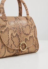 Brunormig väskösa i skinn med glansig yta, rundad topp, strukturerad form och en guldfärgad cirkulär detalj på framsidan.