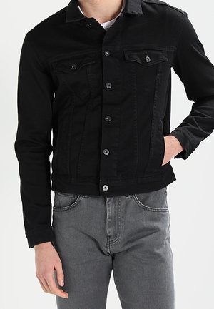 Homme portant une veste en jean noire cintrée avec des boutons et des poches poitrine, associée à un jean gris, main dans la poche.