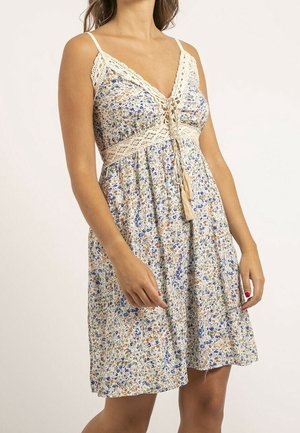 Femme portant une robe d'été fleurie sans manches avec bordure en dentelle, un col en V, et un détail noué à la taille, debout devant un fond uni.