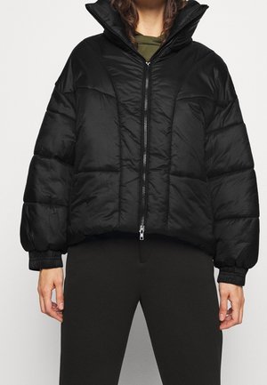 Henkilö pukeutunut mustaan toppatakkiin, jossa on etuvetoketju, korkea kaulus ja joustavat hihansuut, yhdistettynä mustiin housuihin ja oliivinvihreään toppiin alla.