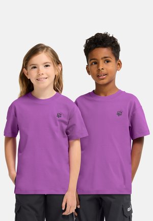 Zwei Kinder tragen identische lila T-Shirts mit kleinen schwarzen Pfotenabdrücken auf der Brust und stehen nebeneinander vor einem schlichten Hintergrund.