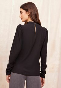 Blusa nera a maniche lunghe con texture a costine, collo alto e dettaglio a chiave sul retro chiuso con tre bottoni.