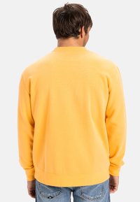 Sudadera amarilla de manga larga, con puños acanalados y cuello redondo, hecha de una suave mezcla de algodón, mostrada desde atrás.