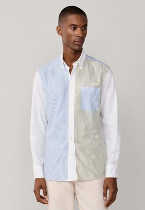Homme portant une chemise à manches longues boutonnée avec des rayures verticales bleues et beiges et une poche poitrine à rayures bleues, associée à un pantalon beige clair.