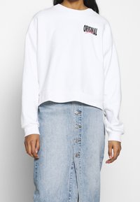 White cropped sweatshirt with "ORIGINAL" graphic in black and pink, paired with a light denim skirt featuring vertical buttons down the front.