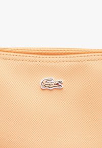 Funda textil de color durazno con cierre de cremallera. Presenta un pequeño logo de cocodrilo bordado en blanco. Superficie texturizada.