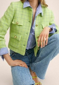 Femme portant une veste texturée vert citron avec des boutons dorés, une chemise rayée bleue, un jean bleu clair et des sandales vert citron, assise en tailleur.