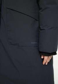 Mörkblå vadderad jacka med en subtil rutad textur, med en sidficka och varumärket "ELVINE" på den nedre delen.