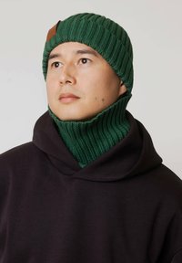 Zielona prążkowana dzianinowa czapka i dopasowana komin, oba wykonane z miękkiego materiału; noszone z czarną bluzą z kapturem; widoczna skórzana naszywka.
