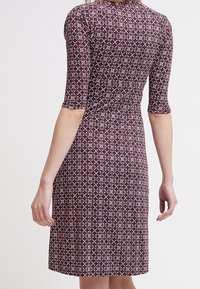 Fitted, kortärmad klänning i mörk lila tyg med ett geometriskt mönster i röd, vit och rosa. Halvlång klippning, slät textur.