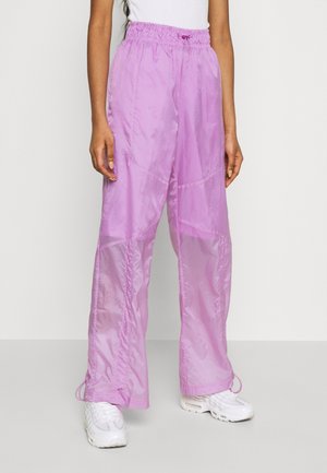 Person wearing loose, sheer lilac pants with elastic waistband and white sneakers, standing against a plain light background.