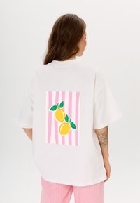 Witte oversized T-shirt met een graphic van twee citroenen met groene bladeren op een roze gestreepte achtergrond. Glad katoenmateriaal.
