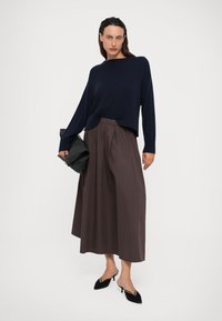 Vrouw in een marineblauwe langemouwen trui, bruine wijde cropped broek, zwarte pumps met een puntige neus, en met een zwarte clutch.