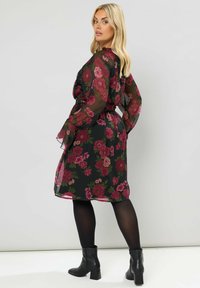 Abito nero floreale con sovrapposizione trasparente, caratterizzato da fiori rosa e verdi, maniche lunghe e una vita cinchata. Indossato con stivaletti neri alla caviglia.