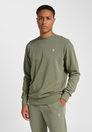 Junger Mann, der einen olivgrünen Pullover und passende Hosen trägt, steht mit einer Hand in der Tasche vor neutralem Hintergrund.