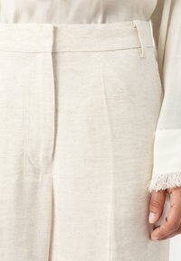 Beige herringbone-kuvioiset housut, joissa on tasainen etuosa, vyölenkit ja kevyt rakenne. Helmassa on raakafrangi-yksityiskohta.