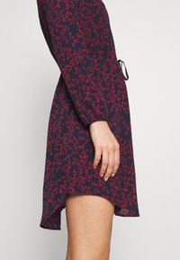 Robe bleu marine à motif floral rouge, avec manches longues, taille cintrée et ourlet incurvé. Le tissu semble léger et texturé.