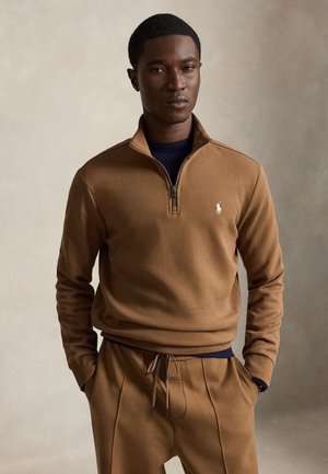 Homme portant un sweat-shirt marron à fermeture éclair sur le quart supérieur et un pantalon assorti, les mains dans les poches, sur un fond neutre uni.