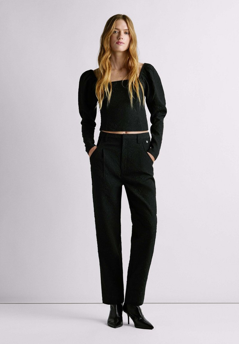 Zwart cropped top van textuurstof, met lange pofmouwen. Zwarte high-waisted broek met diepe plooien, gedragen met puntige zwarte hakken.