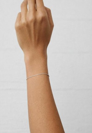 Rechterhand en onderarm opgeheven, met een dunne zilveren kettingarmband, tegen een effen lichtgrijze achtergrond.