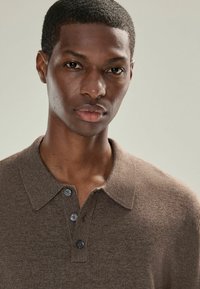 Braunes Strick-Polohemd mit Kragen und drei dunklen Knöpfen. Weicher Stoff und nahtloses Design, getragen von einer Person mit kurzen Haaren.