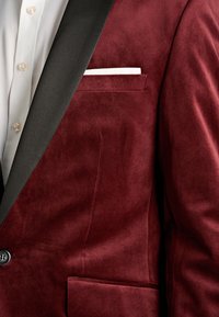 Close-up van een bordeauxrode fluwelen blazer met zwarte revers, enkele zwarte knoop, wit overhemd en wit pochet.