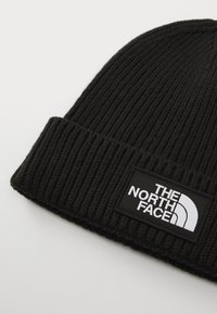 Fekete kötött beanie sapka bordázott textúrával és hajtott szegéllyel. Egy téglalap alakú címke található rajta fehér szöveggel és logóval fekete háttérrel.