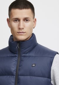 Meripuku takki, jossa on korkea kaulus, vetoketjusuljenta ja logotiedot. Ominaisuuksiin kuuluvat tikkipäällystys, sileä tekstuuri ja istuva muotoilu.