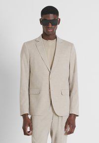 Blazer in lino beige con taglio slim, chiusura a un bottone e due tasche frontali. Abbinato a un top beige coordinato. Occhiali da sole neri.