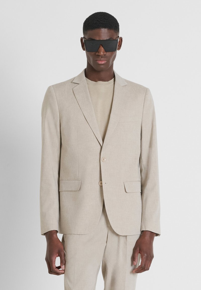 Blazer in lino beige con taglio slim, chiusura a un bottone e due tasche frontali. Abbinato a un top beige coordinato. Occhiali da sole neri.