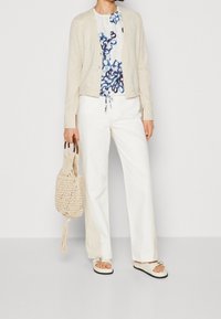 Cardigan beige chiaro, blusa floreale con motivi blu, pantaloni bianchi a gamba larga e borsa intrecciata. Calzature casual con suola spessa.