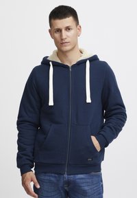 Granatowy hoodie z zamkiem błyskawicznym, w kremowej fleece'owej podszewce, z sznurkami i przednimi kieszeniami. Materiał jest teksturowany, z prążkowanymi mankietami i dołem.