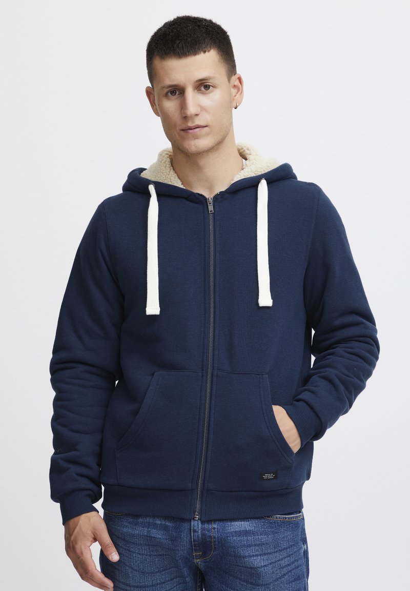 Granatowy hoodie z zamkiem błyskawicznym, w kremowej fleece'owej podszewce, z sznurkami i przednimi kieszeniami. Materiał jest teksturowany, z prążkowanymi mankietami i dołem.
