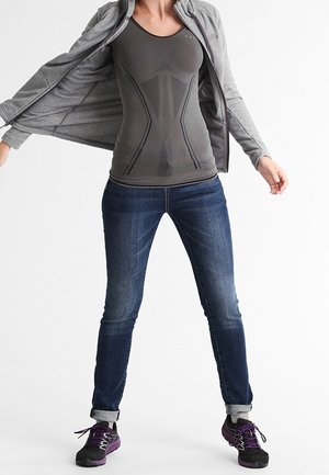 Giacca grisaglia con zip sopra una canotta aderente scura con fantasie testurizzate, jeans blu e sneaker nere con accenti viola.