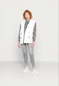Colete branco sem mangas com botões duplos sobre uma blusa de manga longa estampada, combinado com jeans skinny cinzas e tênis brancos.