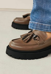 Bruna läderloafers med svart chunky sula, prydda med en dekorativ frans och silverdetalj, bärs med blå denimjeans.