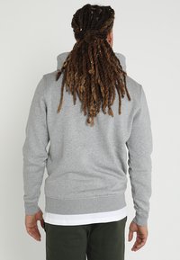 Sudadera gris con cuello alto, puños y dobladillo acanalados. Textura de tela suave; se lleva sobre una camisa blanca, vista desde atrás.