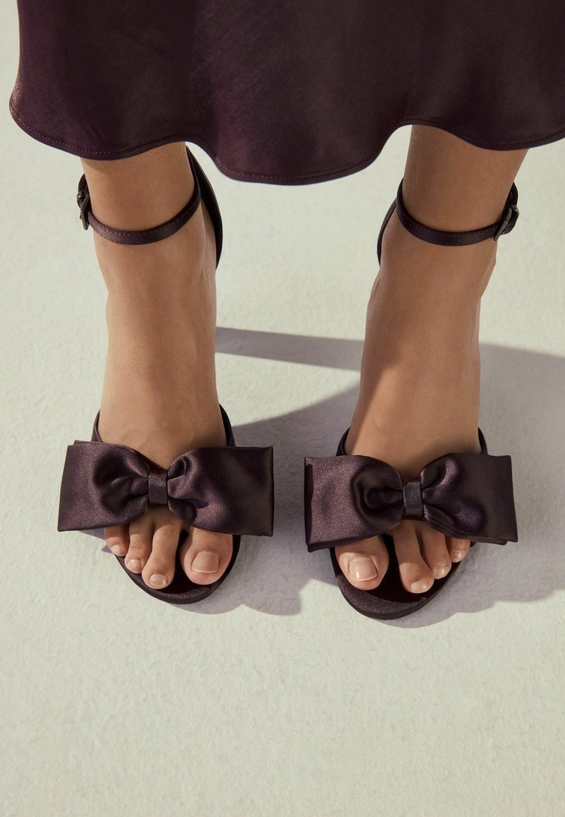 Sandales satinées noires avec un grand nœud décoratif et une bride autour de la cheville. Le design comprend un bout ouvert et une texture lisse.