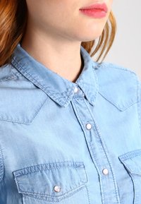 Ljusblå denimskjorta med spetsig krage, framknäppning med knappar och två bröstfickor med rundade silverfärgade knappar.