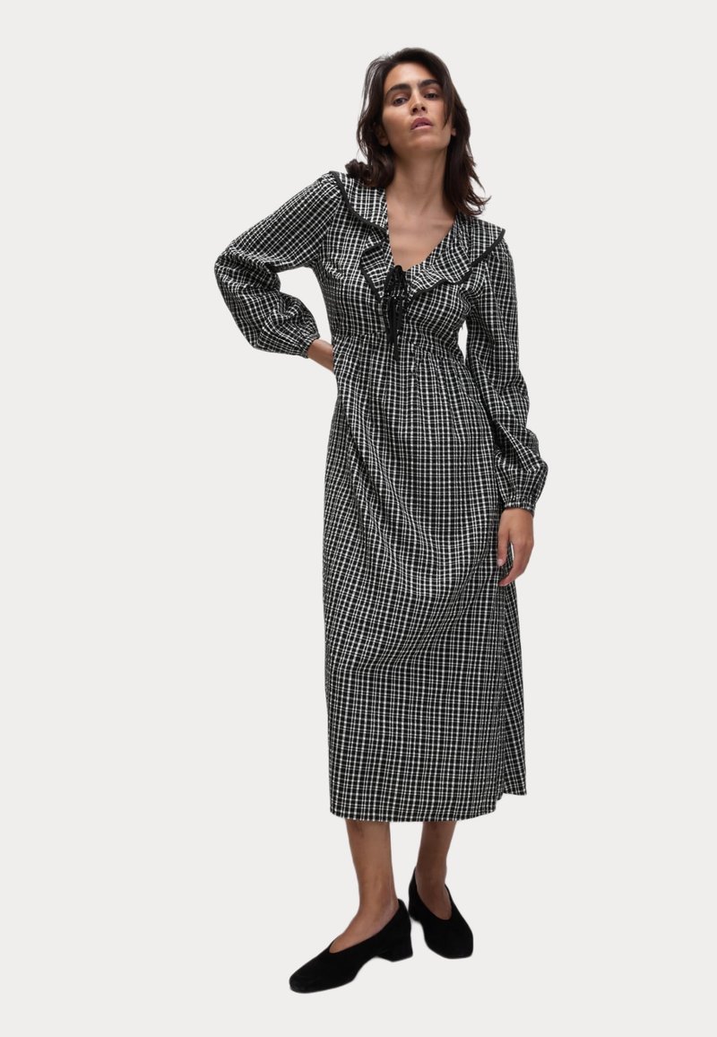 Schwarz-weißer kariierter langer Kleid mit einem Rüschenausschnitt, gebundenem Dekolleté und langen Puffärmeln. Kombiniert mit schwarzen flachen Schuhen.