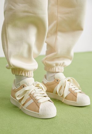 Baskets Adidas beige avec embouts coquille blancs et lacets épais portées avec un pantalon de jogging couleur crème et des chaussettes blanches sur un sol vert.
