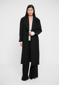 Palton negru din lână cu guler în foma de V, având accesorii aurii la mansete, purtat peste un pulover crem, asortat cu pantaloni negri cu croială largă.