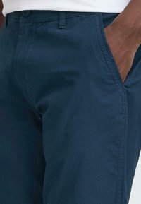 Moški v temno modrih hlačah z desno roko v sprednjem žepu, ki prikazuje teksturo blaga in podrobnosti šivanja.